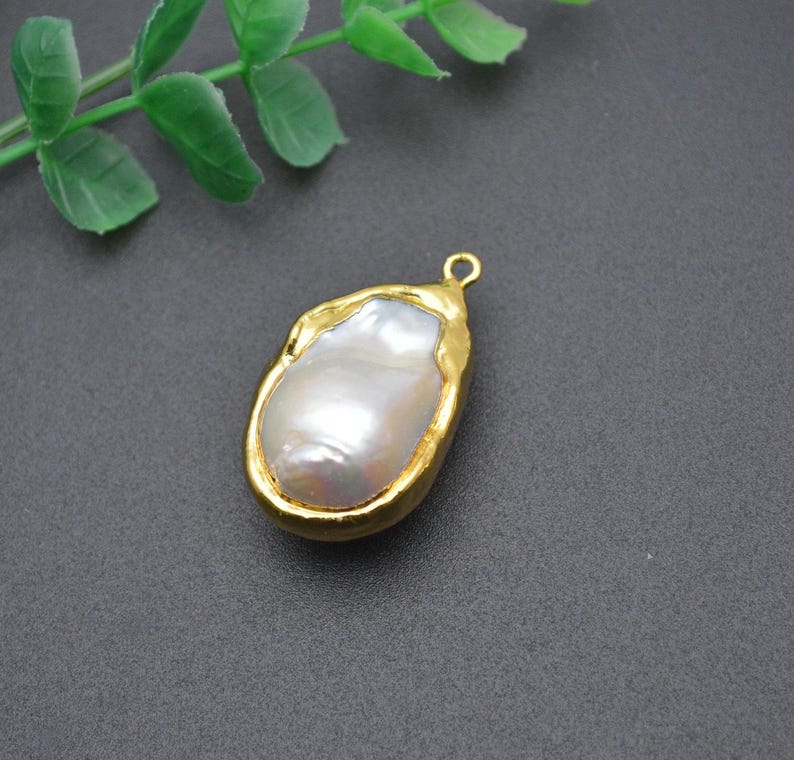 May include: A gold-tone pendant featuring a large, irregular-shaped white pearl. The pearl is set in a gold-tone frame with a textured finish.