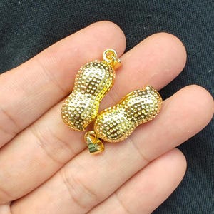 May include: Two gold-colored peanut-shaped charms with a textured surface. Each charm has a loop at the top for attaching to a necklace or bracelet.
