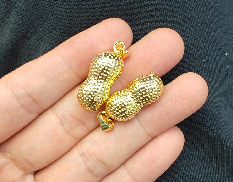 May include: Two gold-colored peanut-shaped charms with a textured surface. Each charm has a loop at the top for attaching to a necklace or bracelet.