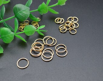 May include: A close-up of two piles of gold jump rings. The jump rings are small and round, and they are arranged in a random pattern. The jump rings are on a black surface.