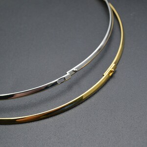May include: Two minimalist necklaces, one silver-colored and one gold-colored, are displayed against a dark background. The necklaces are thin, circular, and have a clasp closure. The gold necklace is positioned below the silver one.