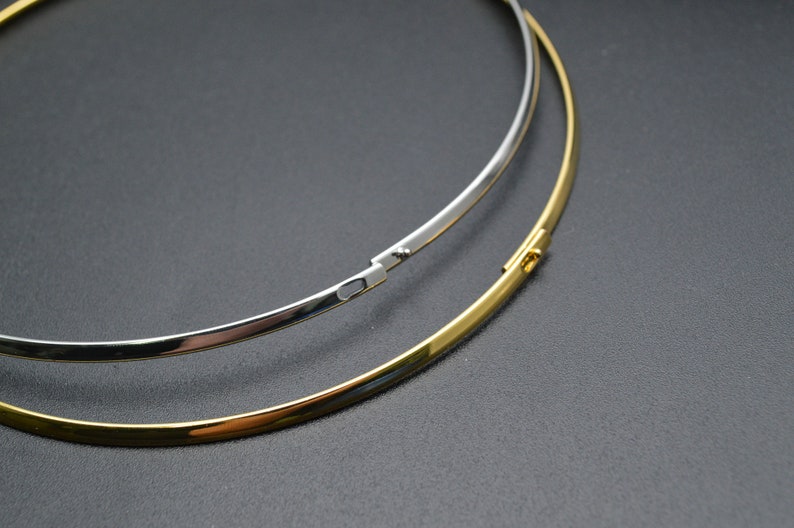 May include: Two minimalist necklaces, one silver-colored and one gold-colored, are displayed against a dark background. The necklaces are thin, circular, and have a clasp closure. The gold necklace is positioned below the silver one.