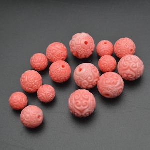 May include: A collection of 15 pink round beads with intricate carved designs. The beads are made of a smooth, polished material and have a slightly glossy finish.