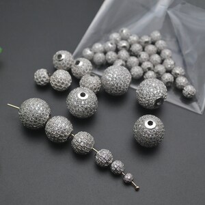 May include: A close-up of a collection of silver round beads with clear rhinestones. The beads are in a clear plastic bag and on a gray surface.