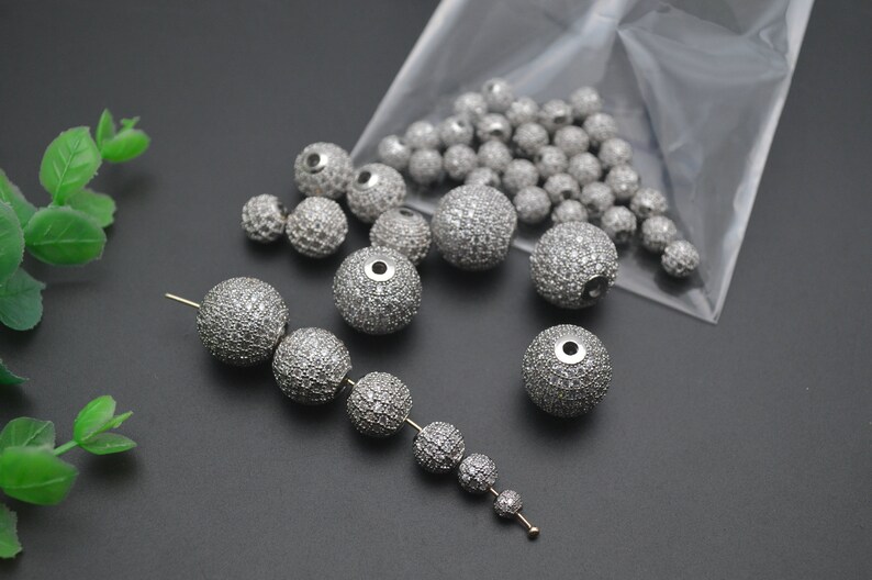 May include: A close-up of a collection of silver round beads with clear rhinestones. The beads are in a clear plastic bag and on a gray surface.