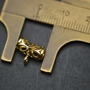 May include: A small, gold-colored jewelry finding with an openwork design featuring a bow shape. The finding is next to a brass ruler, which is used to measure the item. The background is a dark gray surface.