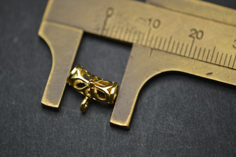 May include: A small, gold-colored jewelry finding with an openwork design featuring a bow shape. The finding is next to a brass ruler, which is used to measure the item. The background is a dark gray surface.