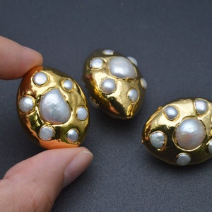 Puede incluir: Tres pendientes ovalados de tono dorado con una perla blanca grande en el centro rodeada de varias perlas blancas más pequeñas incrustadas en la superficie de metal pulido. Los pendientes tienen un acabado liso y reflectante con forma orgánica.