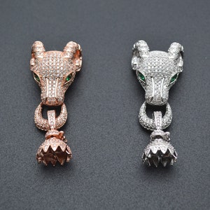 Könnte beinhalten: Zwei silberne und roségoldene Tierkopf-Anhänger mit grünen Augen und klaren Kristall-Akzenten. Die Anhänger sind mit einer strukturierten Oberfläche und einem baumelnden, blumenförmigen Boden verziert.