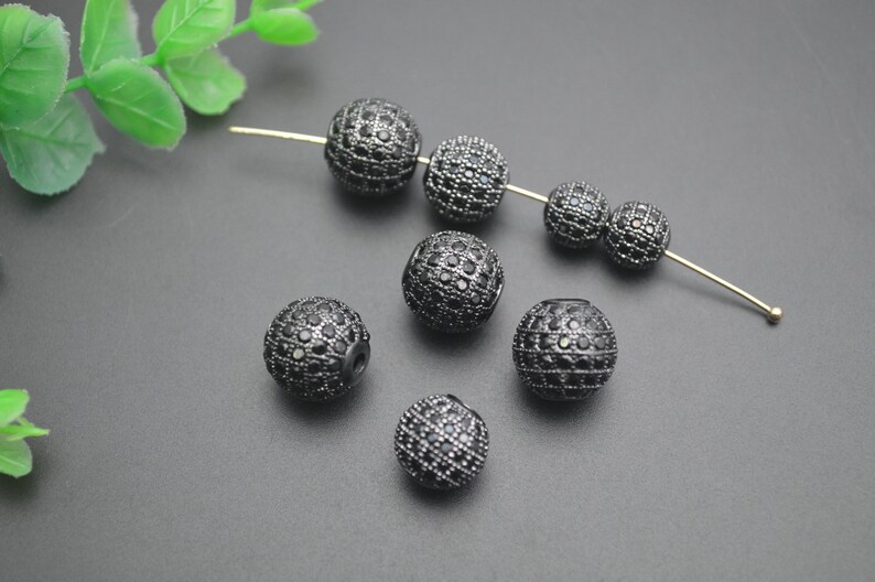 May include: Black round beads with small black rhinestones covering the surface. The beads are scattered on a gray surface.