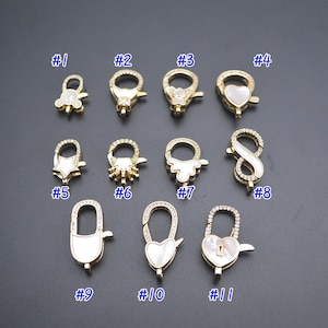 Könnte beinhalten: Elf goldfarbene Metallverschlüsse mit weißen Akzenten. Jeder Verschluss ist von 1 bis 11 nummeriert. Die Verschlüsse sind in verschiedenen Formen gestaltet, darunter Herzen, Sterne und Unendlichkeitszeichen.