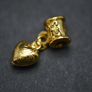 Puede incluir: Un charm dorado con un colgante en forma de coraz&oacute;n y una cuenta cil&iacute;ndrica con detalles decorativos.