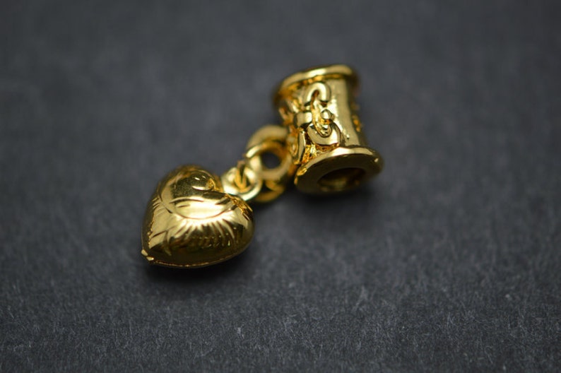 Puede incluir: Un charm dorado con un colgante en forma de coraz&oacute;n y una cuenta cil&iacute;ndrica con detalles decorativos.
