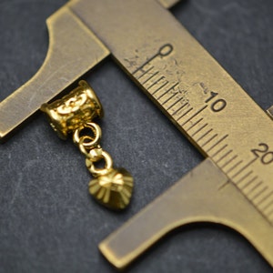 Puede incluir: Pendiente de metal dorado con un dije en forma de coraz&oacute;n colgando de una cadena. El pendiente se est&aacute; midiendo con una regla, mostrando que mide aproximadamente 2,5 cm de largo.