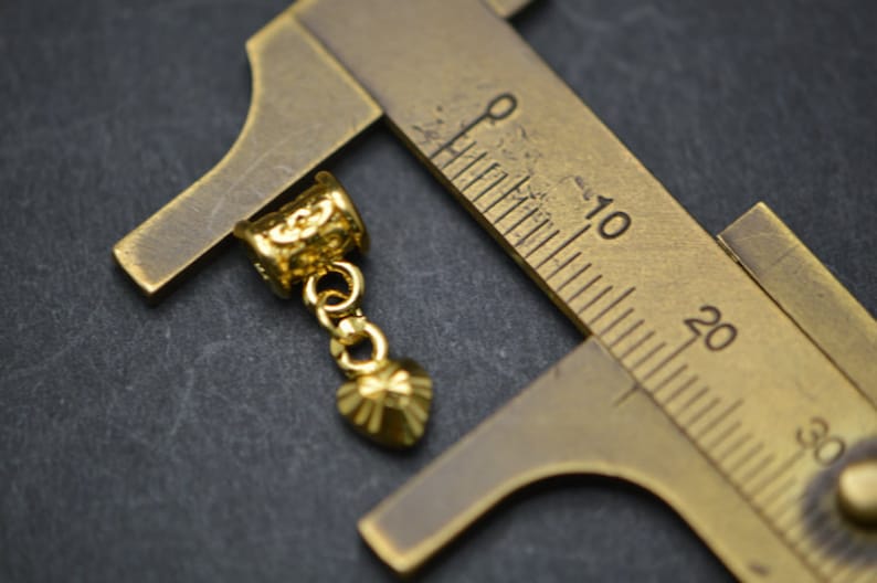 Puede incluir: Pendiente de metal dorado con un dije en forma de coraz&oacute;n colgando de una cadena. El pendiente se est&aacute; midiendo con una regla, mostrando que mide aproximadamente 2,5 cm de largo.