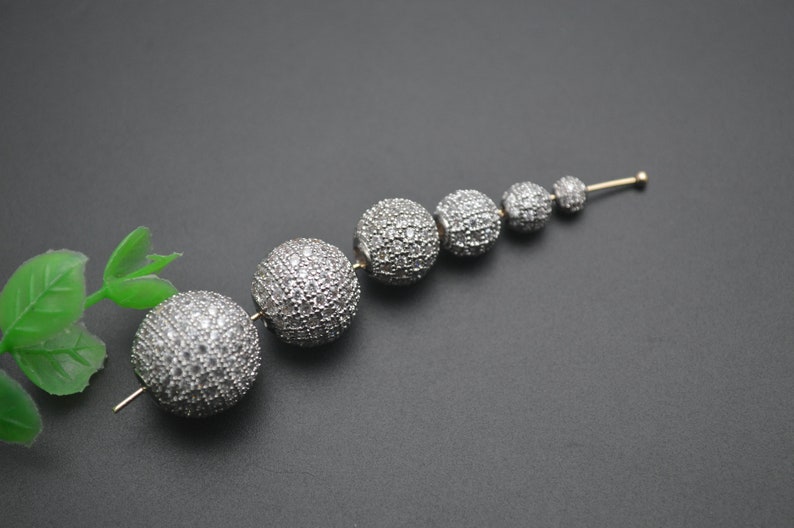 May include: A set of six silver-toned round beads, each covered in small, clear rhinestones. The beads are arranged in a line, decreasing in size from left to right.