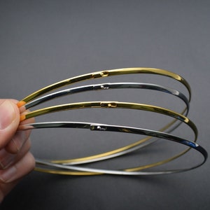 May include: A collection of thin, curved metal chokers in gold and silver tones. Each choker has a clasp closure. The jewelry is held against a dark gray background.