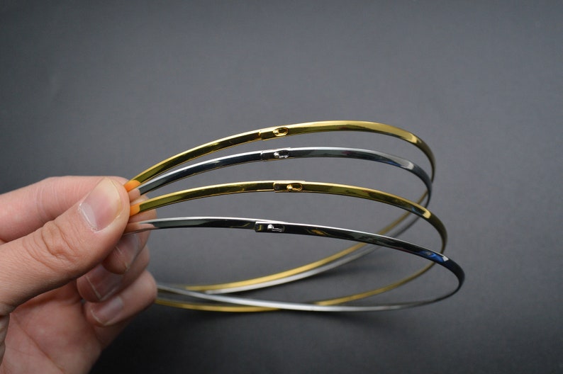 May include: A collection of thin, curved metal chokers in gold and silver tones. Each choker has a clasp closure. The jewelry is held against a dark gray background.