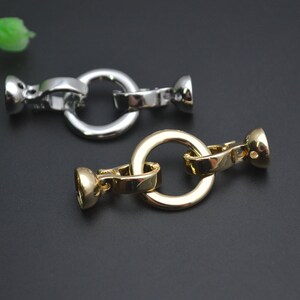 May include: Close-up of two metal jewellery clasps. One is silver-coloured, and the other is gold-coloured. Each clasp has a circular ring and two end pieces. The clasps are on a dark background.