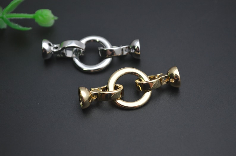 May include: Close-up of two metal jewellery clasps. One is silver-coloured, and the other is gold-coloured. Each clasp has a circular ring and two end pieces. The clasps are on a dark background.
