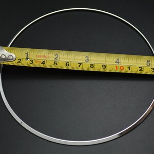 May include: A silver-colored necklace with a circular design, measured with a yellow tape measure. The necklace appears to be approximately 6 inches in diameter. The background is a solid black.