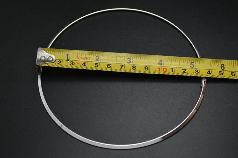 May include: A silver-colored necklace with a circular design, measured with a yellow tape measure. The necklace appears to be approximately 6 inches in diameter. The background is a solid black.