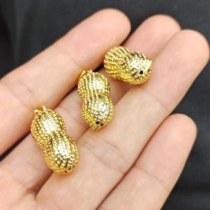 May include: Three gold-colored peanut-shaped charms with a textured surface. The charms are small and have a hole at the top for attaching to a necklace or bracelet.