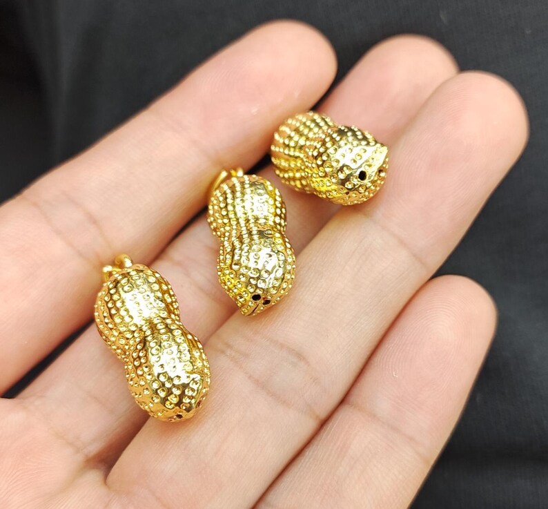 May include: Three gold-colored peanut-shaped charms with a textured surface. The charms are small and have a hole at the top for attaching to a necklace or bracelet.