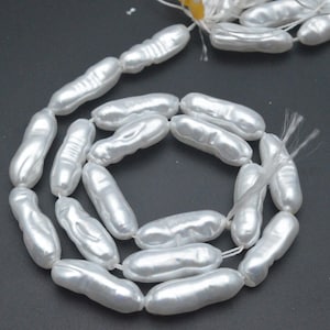 May include: A strand of white, oblong-shaped pearls. The pearls are strung together on a white string, and are arranged in a circular pattern against a dark background. The pearls have a lustrous, pearlescent sheen.