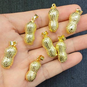May include: Six gold-colored peanut-shaped pendants with a textured surface. The pendants are on a hand.