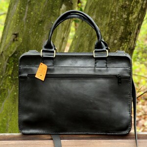 May include: A black leather briefcase with a top handle and a shoulder strap. The bag has a zippered front pocket and gold-colored hardware. A small tag hangs from the zipper. The background is a blurred view of a park.