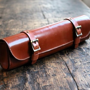 May include: A brown leather roll-up case with two buckled straps. The case is rectangular with rounded ends and a glossy finish. It is resting on a weathered wooden surface.