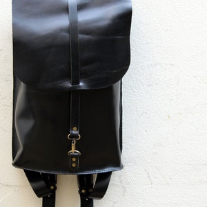 May include: A black leather backpack with a flap closure and adjustable straps. The bag has a simple design with a single strap closure and brass-coloured hardware. The backpack is hanging against a white wall.