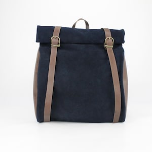 Puede incluir: Una mochila de lona azul oscuro con correas y hebillas de cuero marrón. La mochila tiene un cierre de rollo.