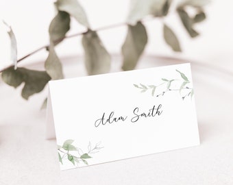May include: White place card with a green leafy design and the name "Adam Smith" written in black cursive.