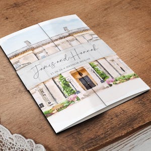 May include: A folded wedding invitation featuring a watercolor illustration of a stately building. The invitation is white with the names "James and Hannah" and the date "27.06.26" printed on the front. The invitation is on a wooden surface.