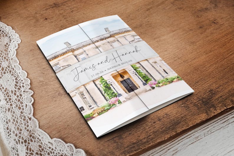 May include: A folded wedding invitation featuring a watercolor illustration of a stately building. The invitation is white with the names "James and Hannah" and the date "27.06.26" printed on the front. The invitation is on a wooden surface.