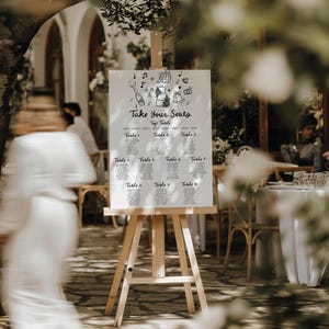 May include: A white wedding seating chart with black doodle illustrations of a cake, champagne bottle, music notes, and a disco ball. The chart is titled "Take Your Seats" and lists table numbers 1 through 10. The chart is displayed on a wooden easel.
