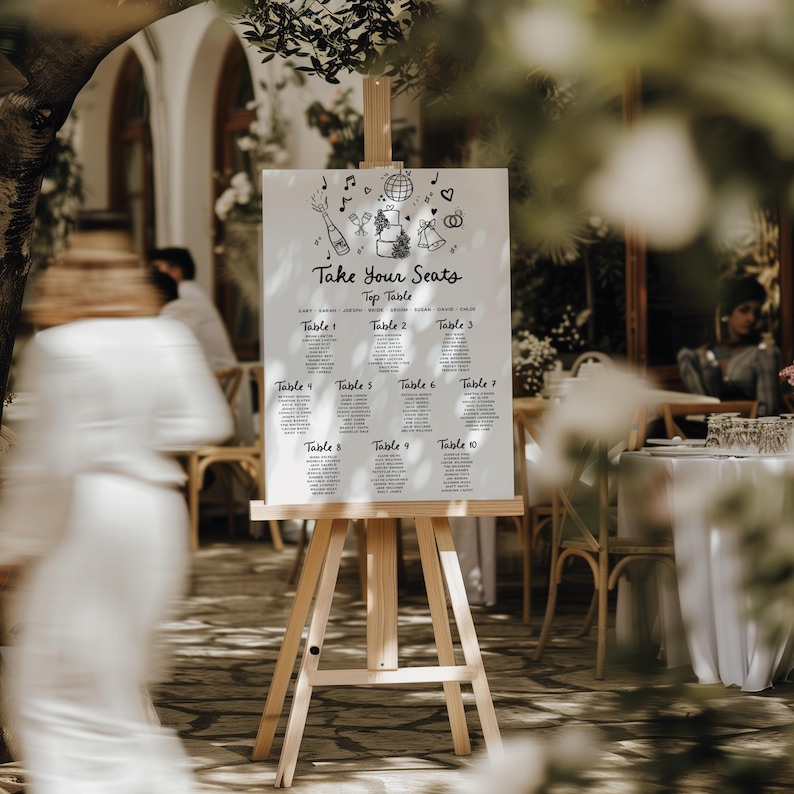 May include: A white wedding seating chart with black doodle illustrations of a cake, champagne bottle, music notes, and a disco ball. The chart is titled "Take Your Seats" and lists table numbers 1 through 10. The chart is displayed on a wooden easel.