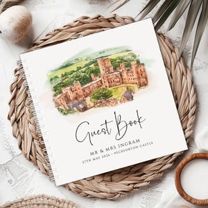May include: A spiral-bound guest book with a watercolor illustration of a castle. The cover reads "Guest Book" in elegant script, with the names "MR & MRS INGRAM" and the date "27TH MAY 2026" below the title. The book rests on a woven mat.