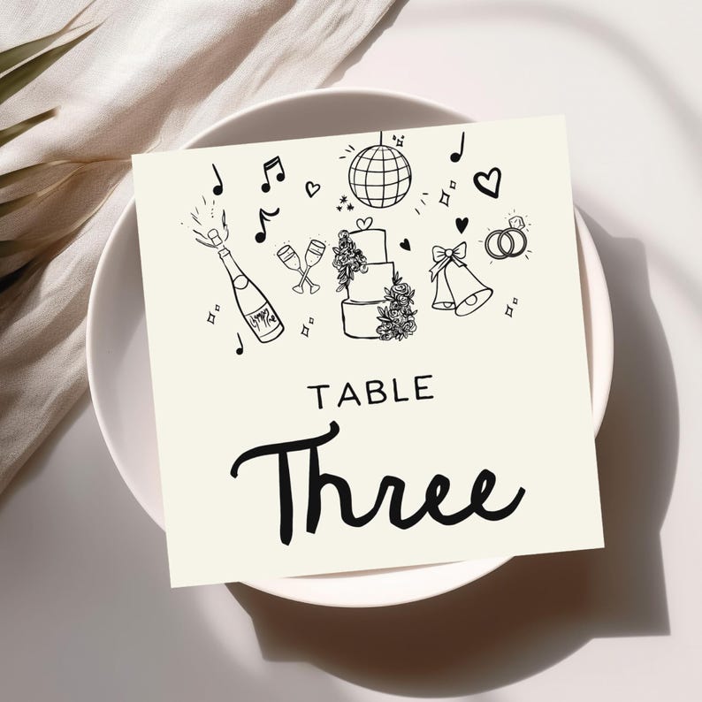 May include: A white table number card with black illustrations of a wedding cake, champagne flutes, music notes, a disco ball, wedding rings, and bells. The text "TABLE Three" is written in black cursive font.