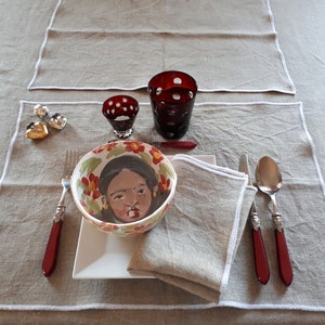 Peut inclure: Un couvert de table avec une assiette blanche, un bol avec un visage peint, une serviette rouge et des couverts rouges et argentés. Il y a deux bougeoirs en verre rouge et deux ornements en forme de cœur argentés sur la table.
