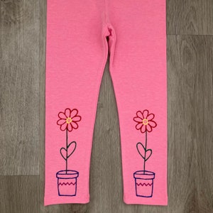 May include: Pink leggings with embroidered flower pot and flower designs. The flowers have red petals and yellow centers. The flower pots are blue. The leggings are made of a soft material.