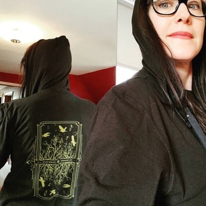May include: Black hooded sweatshirt with a gold graphic on the back. The graphic features a tree with branches and birds, surrounded by a square frame with geometric designs. The text "BLACK SWAN" is visible at the bottom of the graphic.
