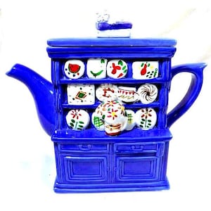 May include: A cobalt blue ceramic teapot shaped like a cabinet, featuring shelves with painted miniature plates and a small figurine on top. The teapot has a curved spout and handle, with two drawers at the base. The plates are decorated with various colorful designs.