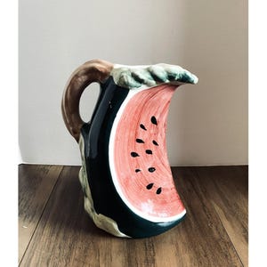 May include: A ceramic pitcher with a watermelon design. The pitcher features a brown handle, a dark green exterior, and a red and black watermelon slice on the front. The rim is decorated with green leaves.