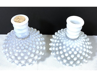 May include: Two white hobnail glass perfume bottles. Each bottle has a round, textured body covered in small, raised dots. The necks are cylindrical, with one bottle having a cork stopper. The bottles are displayed on a white surface.