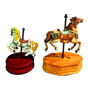 May include: Two carousel horse figurines. One is white with yellow mane and tail, adorned with floral details, atop a red base. The other is tan with a brown mane and tail, embellished with jewels, on a wooden base. Both have a central pole.