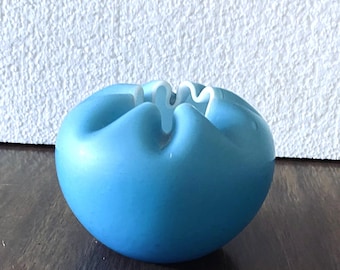 May include: A round, light blue decorative bowl with a ruffled, white-edged opening. The bowl is sitting on a dark wooden surface, with a textured white wall in the background. The bowl's shape is reminiscent of a flower.