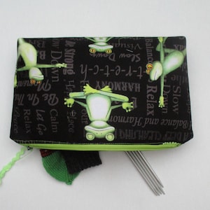 Könnte beinhalten: Schwarze Reißverschlusstasche mit grünem Reißverschluss-Zipper. Die Tasche hat ein grünes Frosch-Design mit den Worten "Slow Down", "Balance", "Relax", "Harmony", "Be In The Moment", "Calm", "Let Go", "Relax", "Peace", "Be Strong", "Happy Place", "Slow Down", "Visual", "Y-t-r-e-t-c-h" darauf gedruckt. Die Tasche ist offen und enthält grüne Stricknadeln.